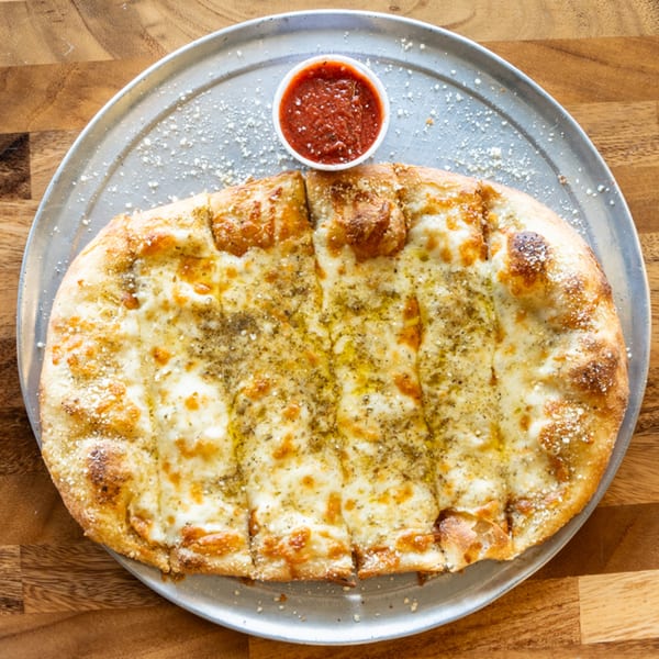 Garlic breadsticks with melted cheese and herbs, served with a side of marinara sauce on a metal tray.