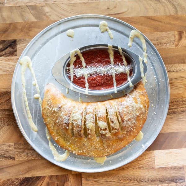 A golden-brown calzone with parmesan cheese and a side of marinara sauce on a metal pizza pan.