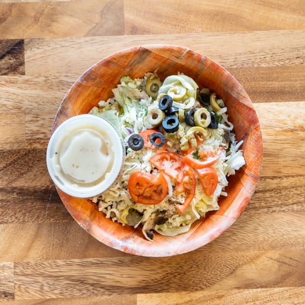 A wooden bowl of salad with lettuce, tomatoes, olives, and a side of dressing on a wooden table.